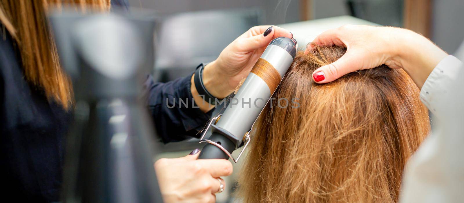 Hands of female hairstylist curls hair client with a curling iron in a hairdressing salon, close up. by okskukuruza