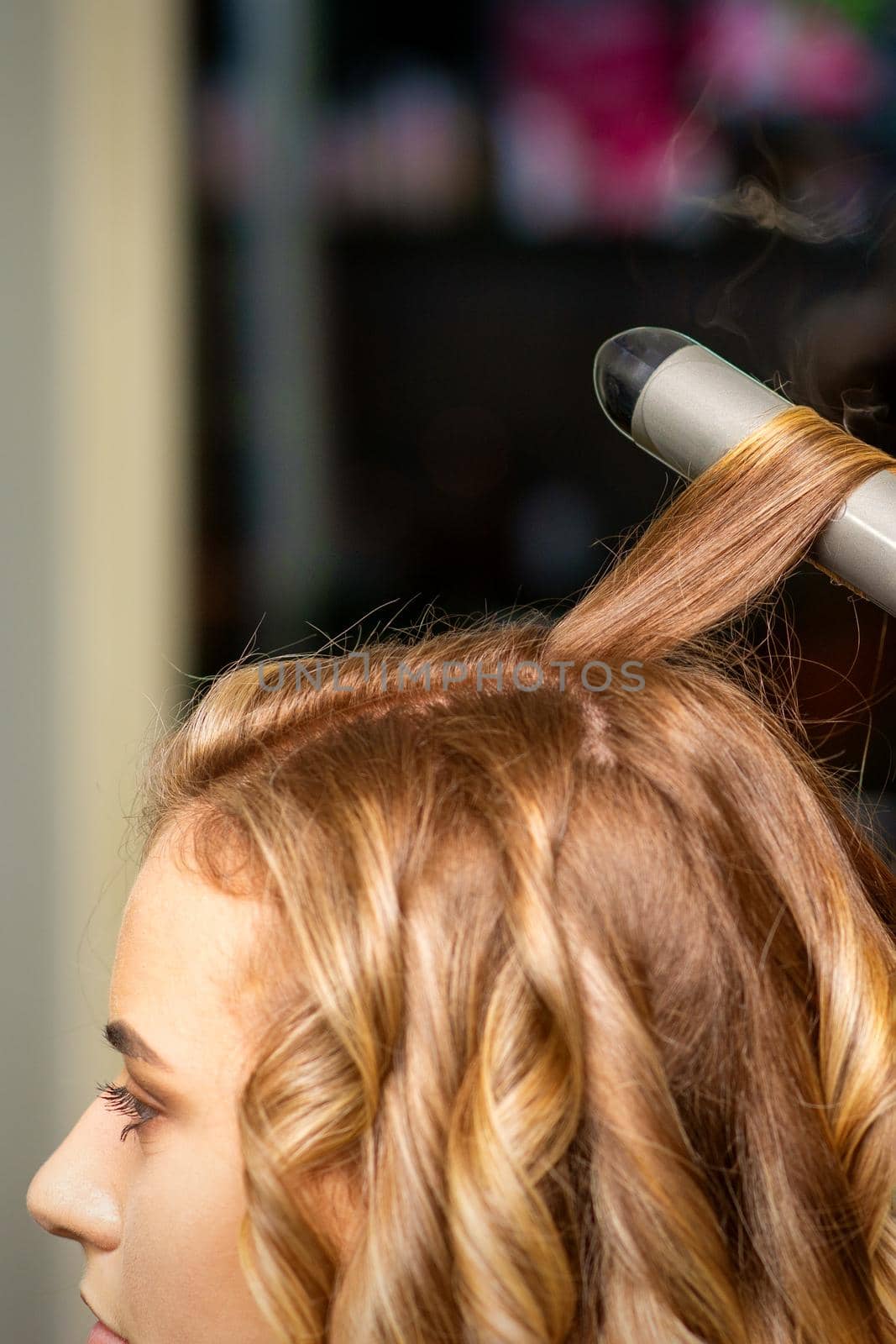 Hairdresser makes curls with a curling iron for the young woman with long brown hair in a beauty salon. by okskukuruza