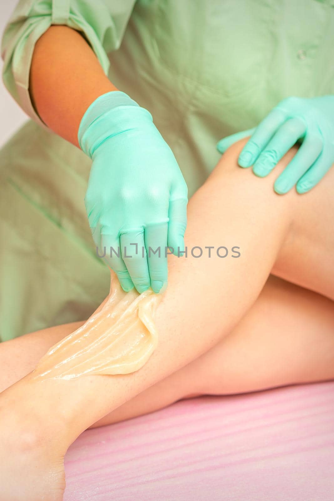 Sugaring legs. Woman legs hair removing. Hands in pink and blue rubber gloves of two beauticians apply sugar paste on female feet. by okskukuruza
