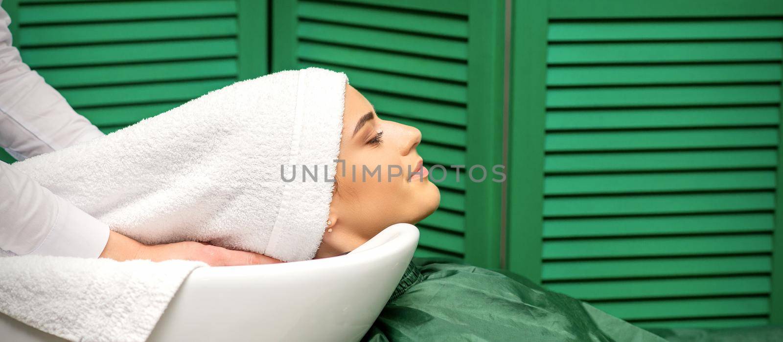 Hairdresser wraps hair of a young caucasian woman in a white towel after washing head in the hairdressing salon. by okskukuruza