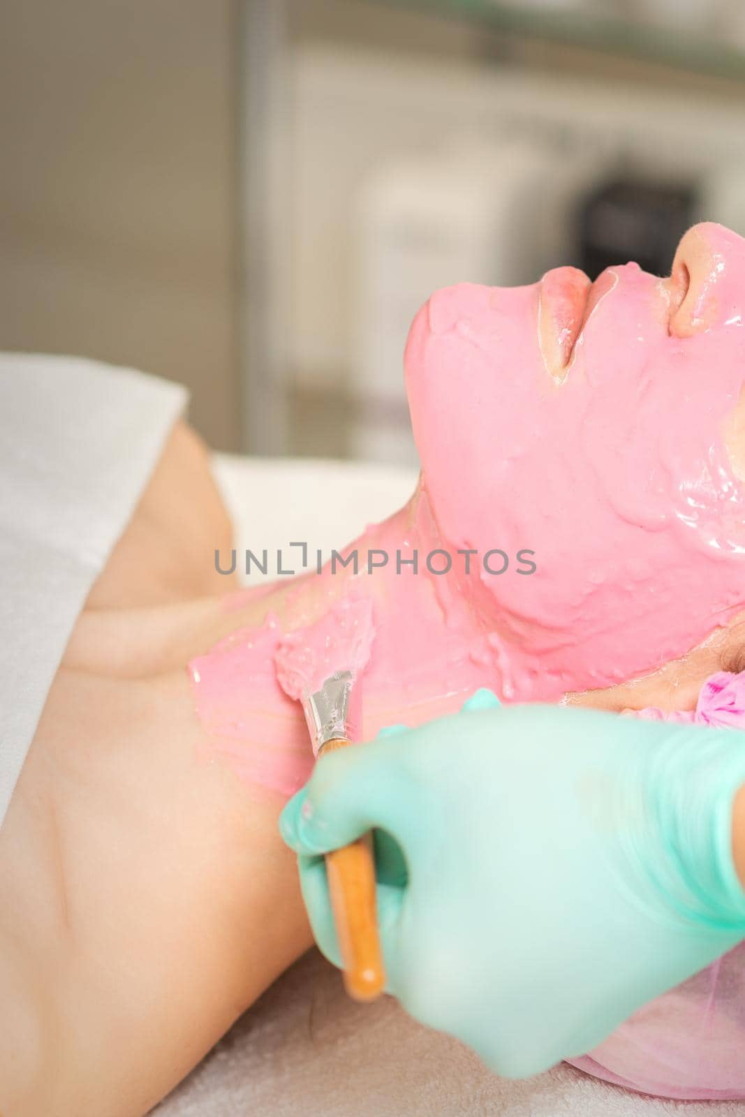 vHand of cosmetologist applying the pink alginic mask with the brush to the face of the young woman in a beauty salon. by okskukuruza