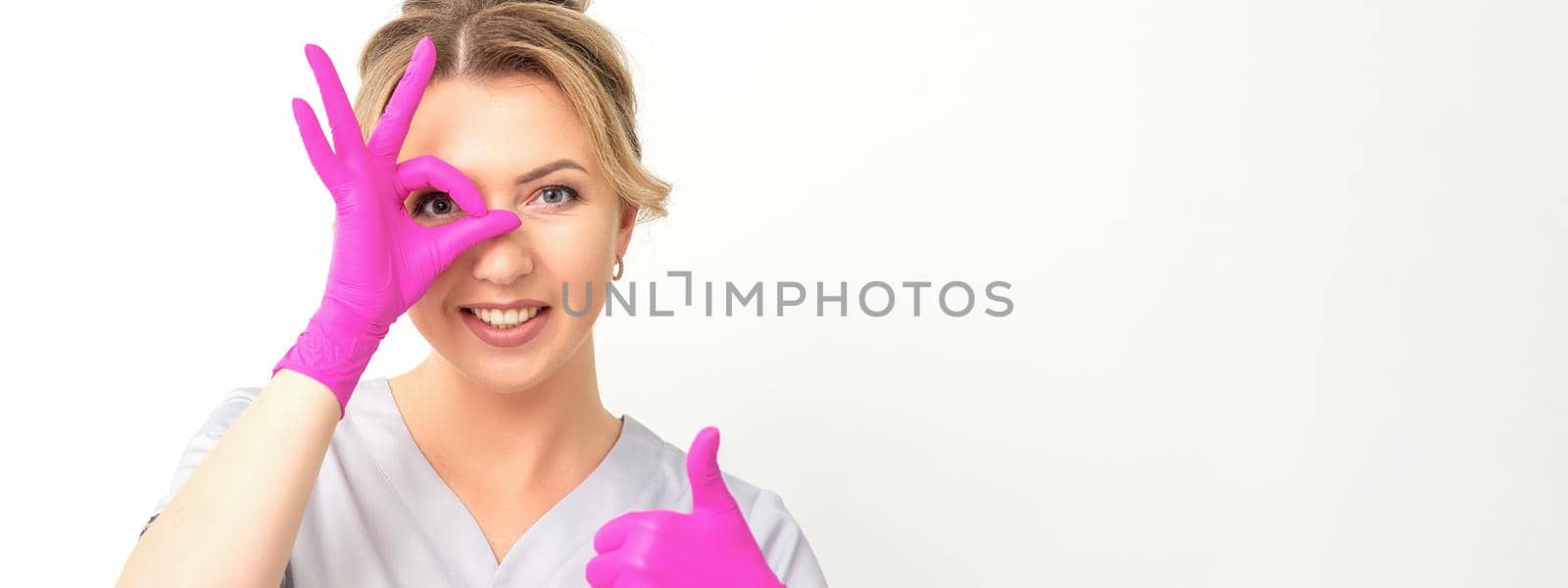 Smiling doctor oculist caucasian woman wearing pink rubber gloves in uniform showing ok sign covering the eye and thumb up gesture against a white background. by okskukuruza