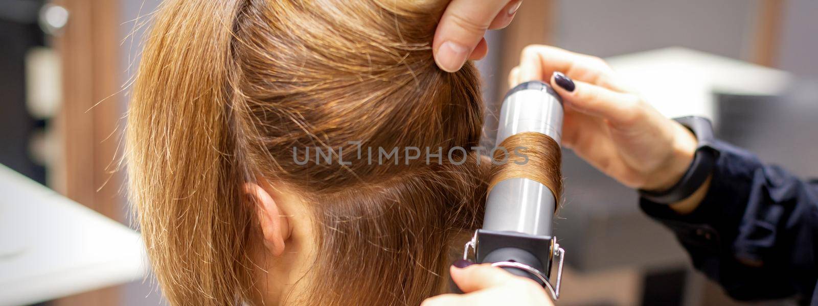 Hands of female hairstylist curls hair client with a curling iron in a hairdressing salon, close up. by okskukuruza