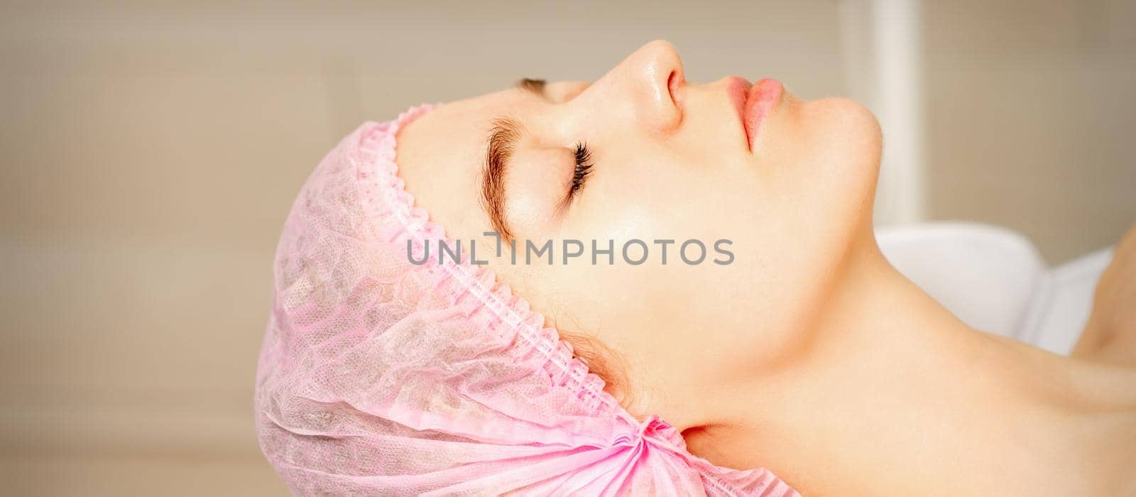 Side view of a young caucasian woman in a medical cap lying with closed eyes waiting for the cosmetic procedure in a beauty salon. by okskukuruza