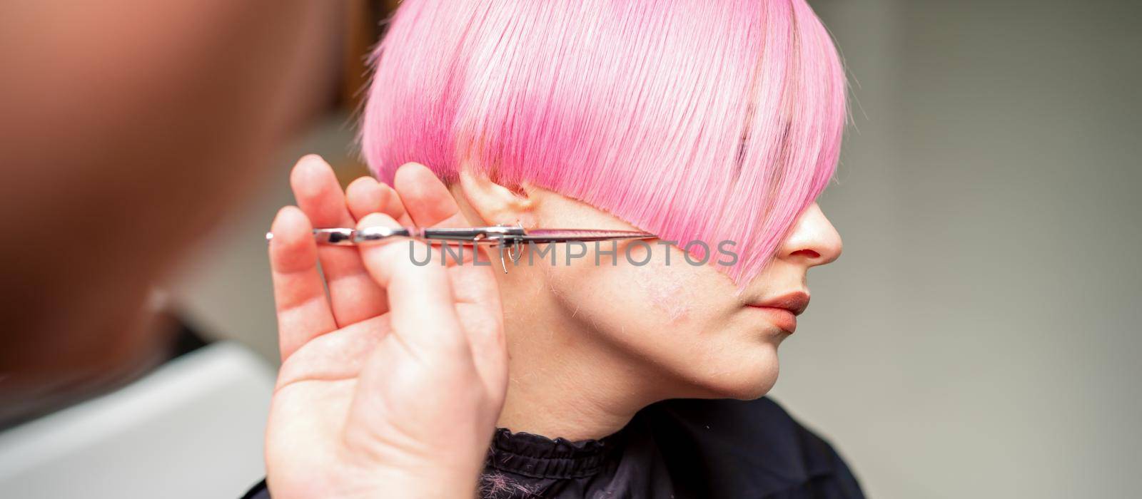 Hand of a hairdresser cutting short pink with scissor hair in a hairdressing salon, close up, side view. by okskukuruza