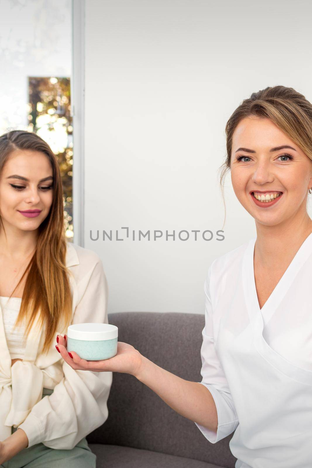 Beautician offering product for young woman, shows thumb up, holding a white plastic jar with a cream, mockup, copy space