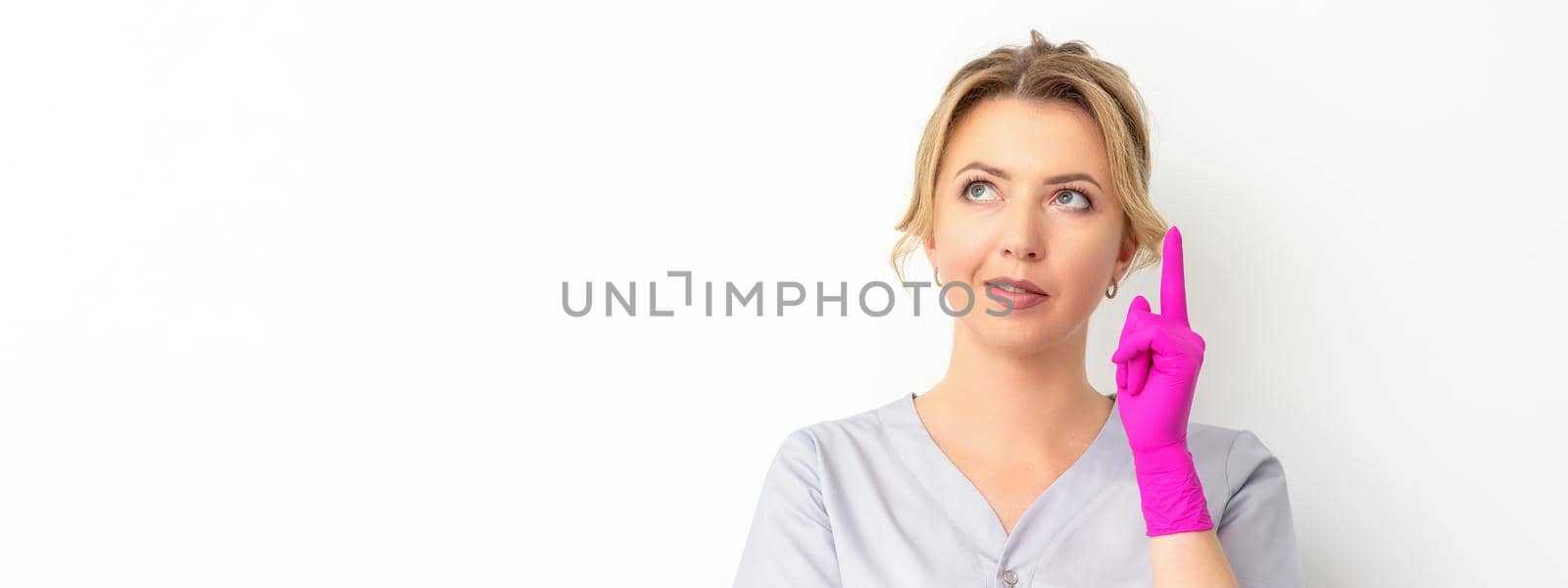 Portrait of young caucasian doctor woman wearing rubber gloves in medical uniform with one finger up, looking up against a white background, copy space. by okskukuruza