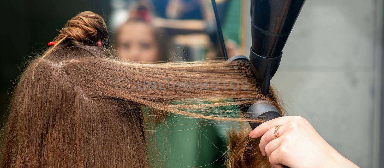 A hairdresser is drying long brown hair with a hairdryer and round brush in a hairdressing salon. by okskukuruza