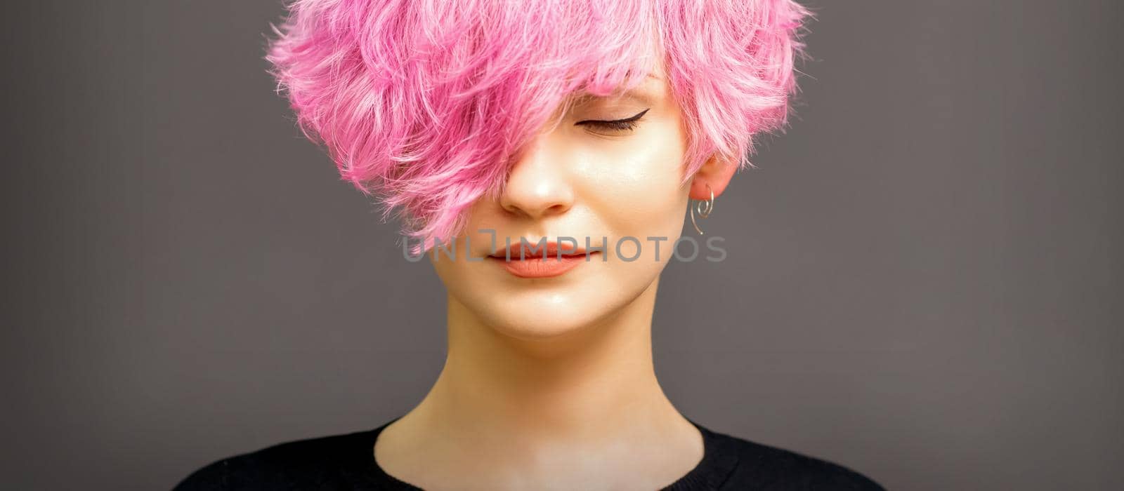 Beautiful young caucasian woman with short curly bob hairstyle dyed in pink color with closed eyes against dark gray background with copy space. by okskukuruza
