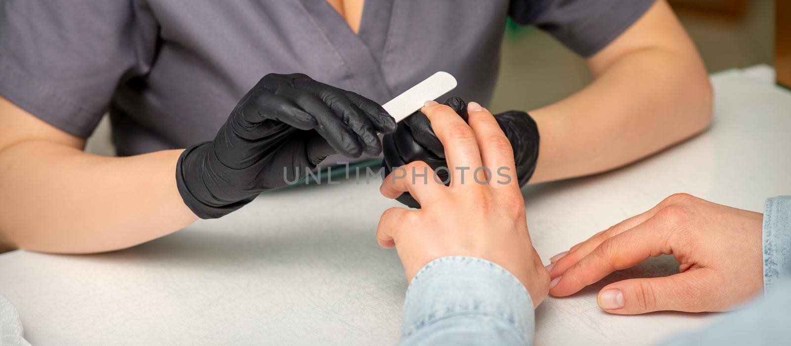 Close up of the caucasian hands of a professional manicurist are filing the nails of a young woman. Young caucasian woman receiving a manicure by a beautician with a nail file in a nail salon. by okskukuruza