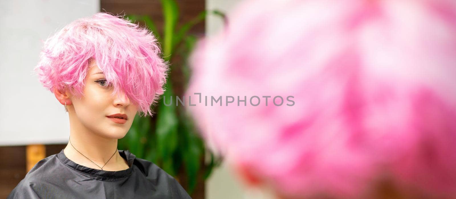 The beautiful young caucasian woman with a new pink short hairstyle in the mirror reflection looking at the camera in a hairdresser salon. by okskukuruza