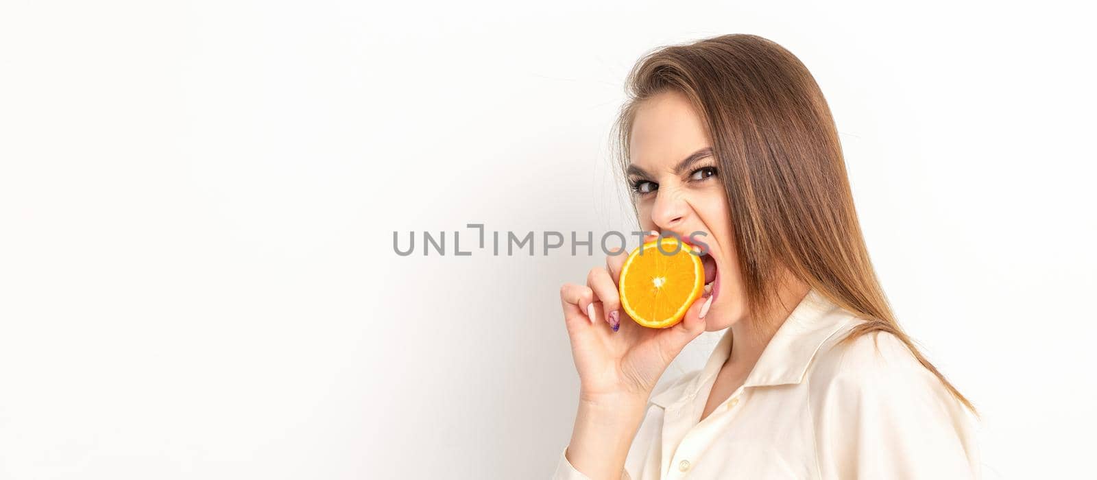 Young caucasian pretty cunning brunette woman biting one orange half and looking at the camera wearing a white shirt over white background. by okskukuruza