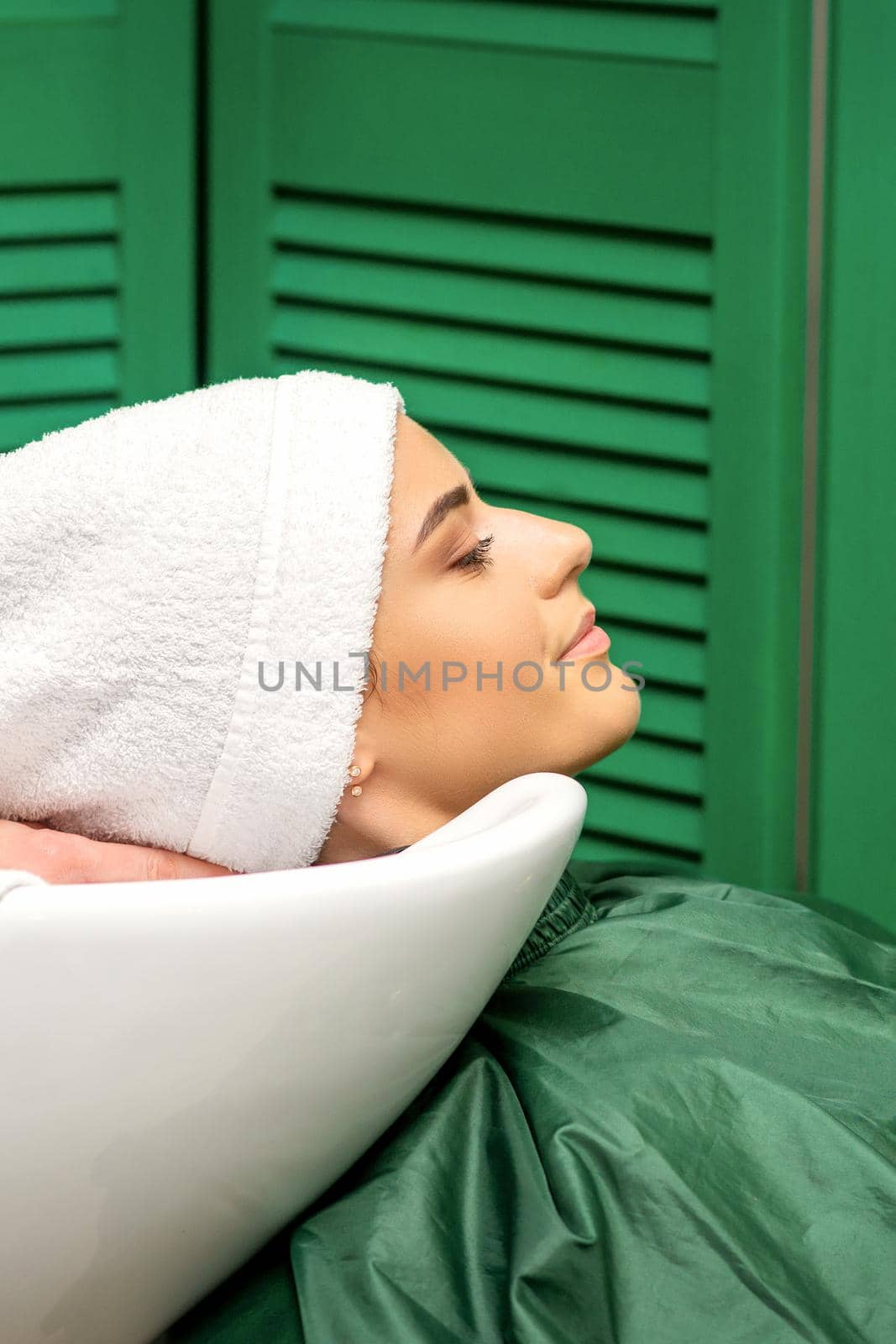 Hairdresser wraps hair of a young caucasian woman in a white towel after washing head in the hairdressing salon. by okskukuruza