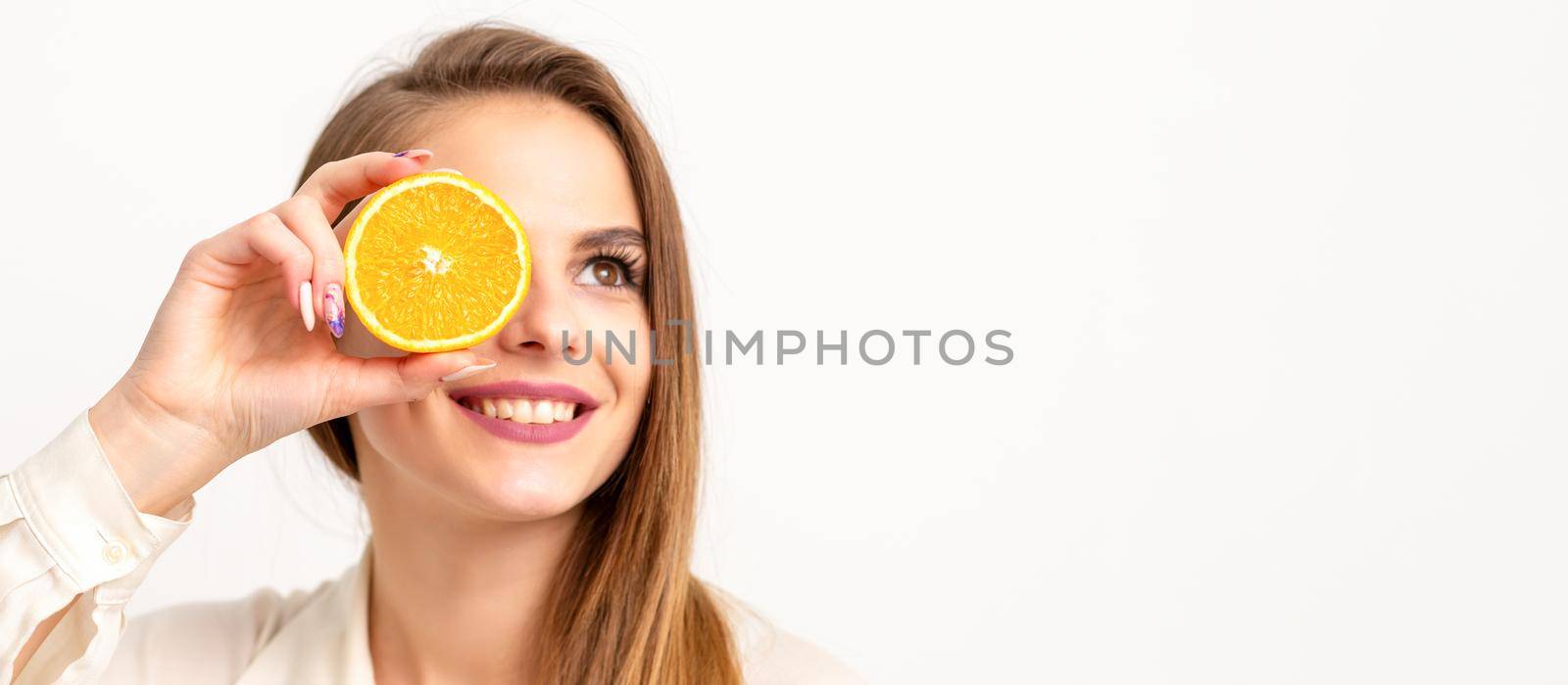 Portrait of a cheerful caucasian young woman covering eye with an orange slice wearing a white shirt over a white background. by okskukuruza