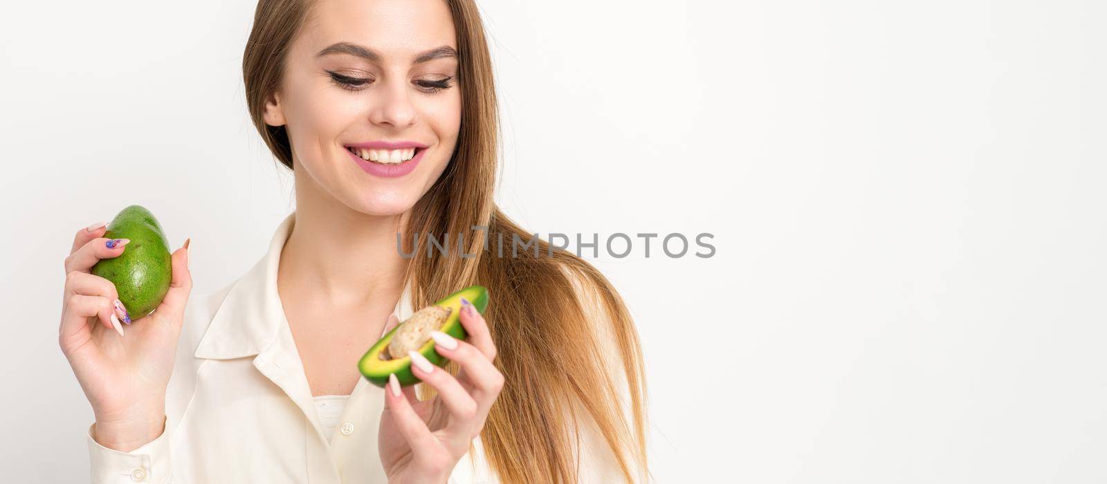 Portrait of a lovely smiling young brunette caucasian woman wearing the white shirt with long hair holding and showing avocado, standing isolated over white background. by okskukuruza