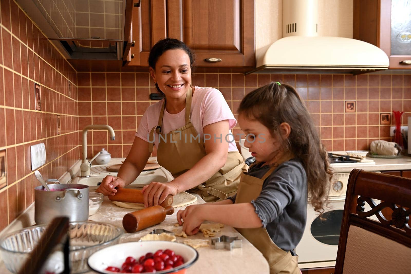 Smiling pretty woman, loving mother using rolling pin, rolls out dough and smiles a beautiful cheerful toothy smile while admiring her cute daughter, helping her to cook delicious festive cherry pie