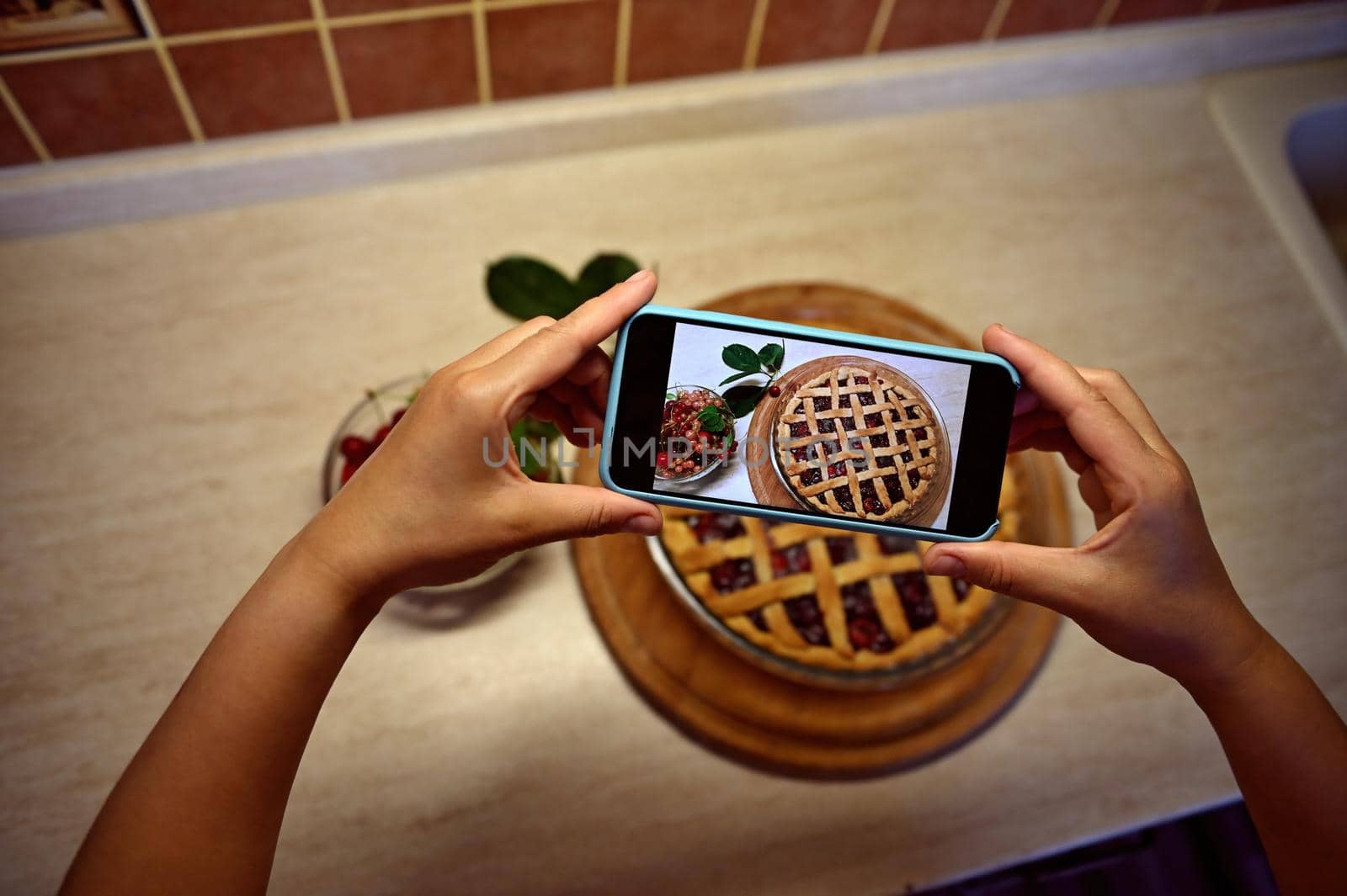 View from above of the hands of a female food blogger holding a smartphone and taking a picture of a classic American puff pastry cherry pie. Mobile phone in live view mode. Baking pastry items