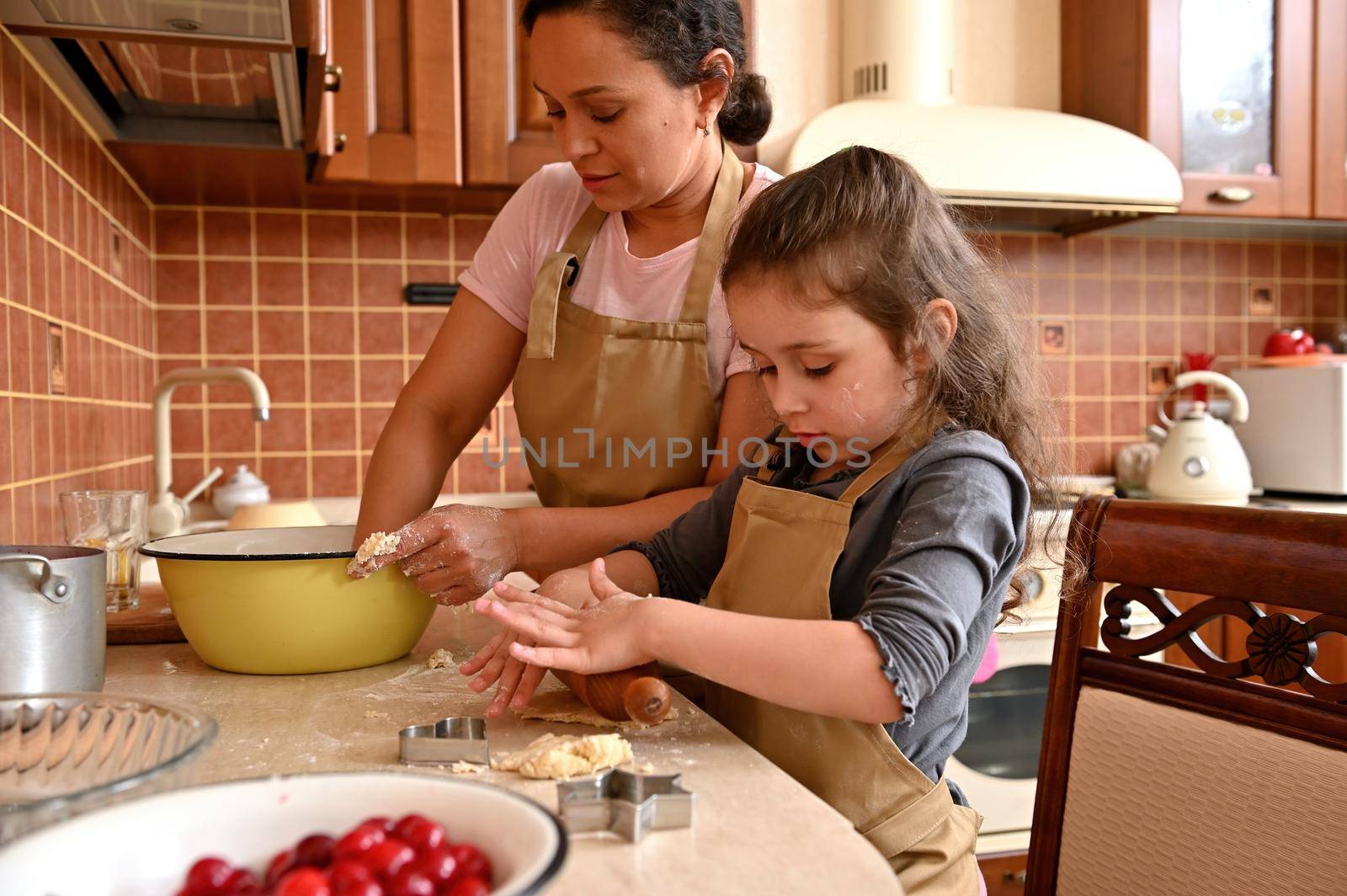 Cute multi-ethnic woman, a loving young mother kneading dough, standing at kitchen table next to her mischievous daughter rolling out the dough, using a wooden rolling pin, preparing pastries and pie