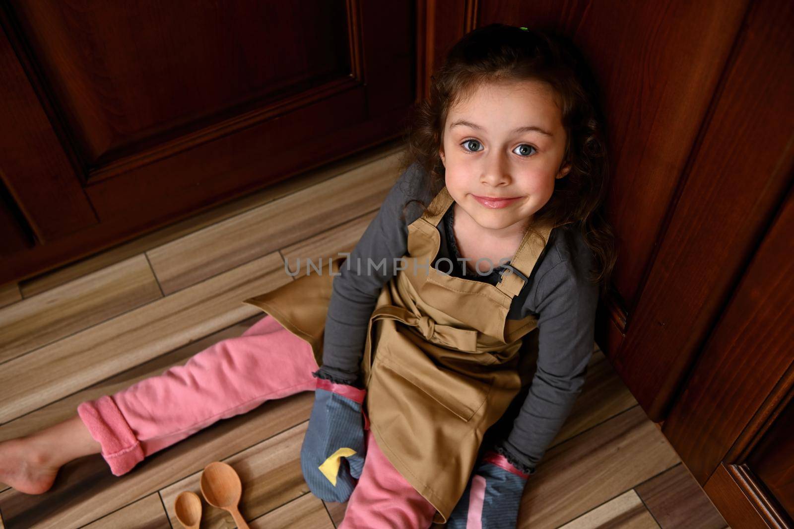 Top view cute child girl, little baker confectioner in a beige chef apron and kitchen mittens sweetly smiling to camera by artgf