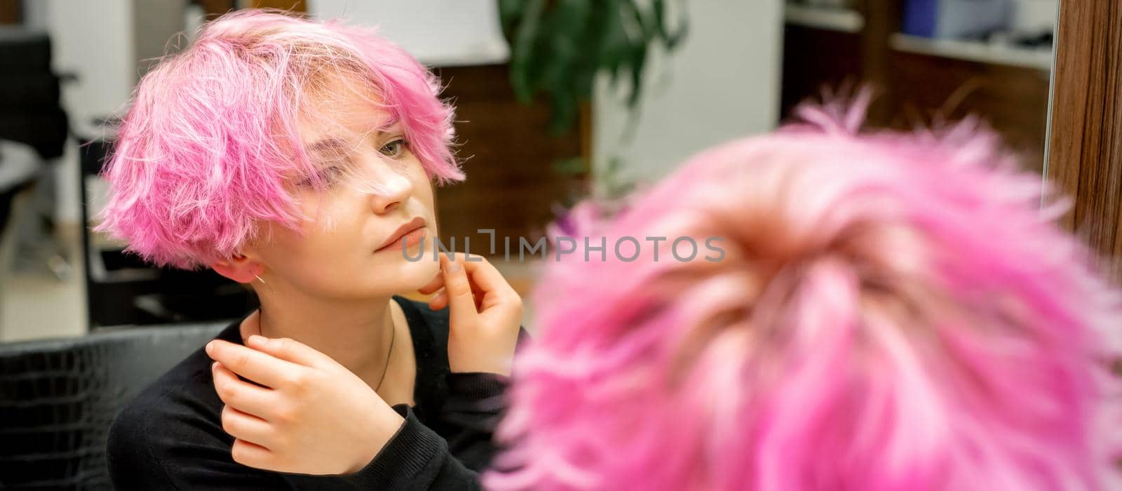 The beautiful young caucasian woman with a new short pink hairstyle looking at her reflection in the mirror checking hairstyle in a hairdresser salon. by okskukuruza