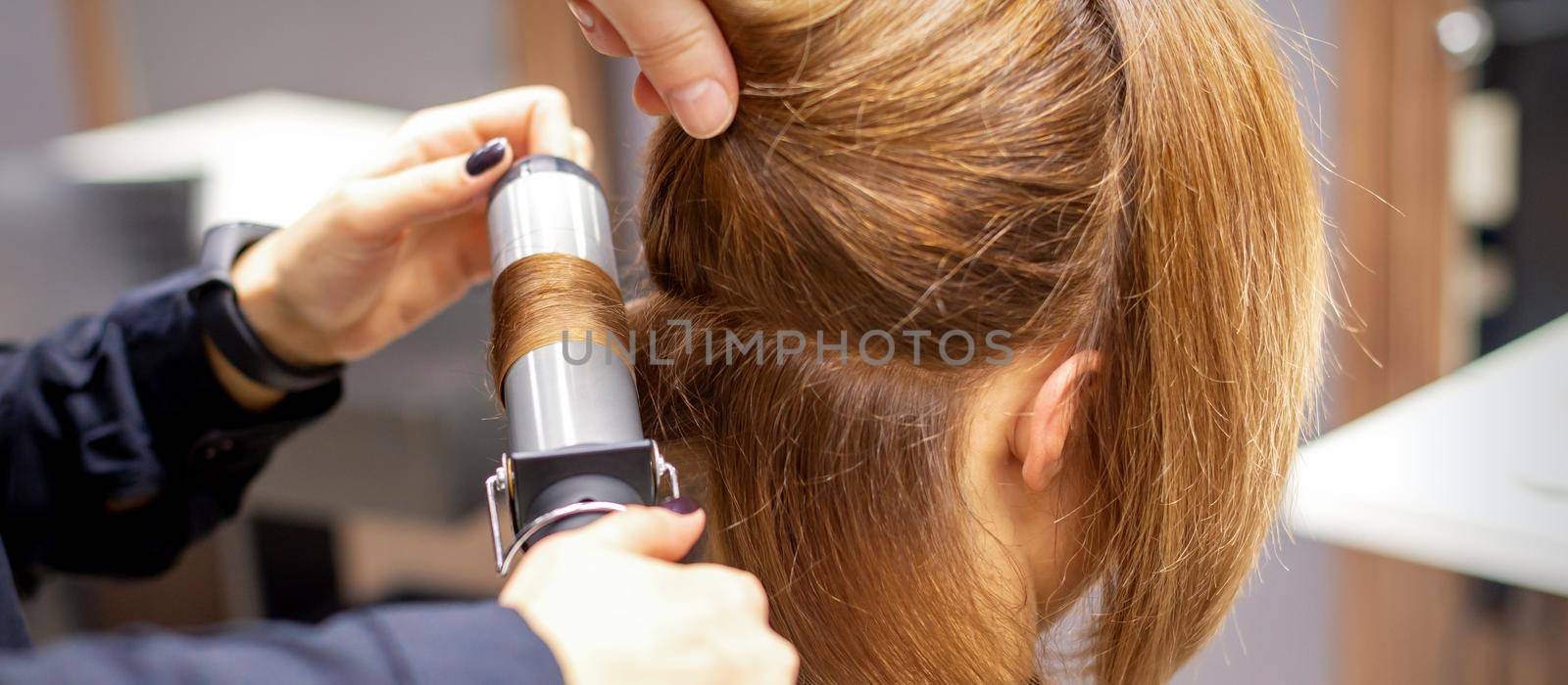 Hands of female hairstylist curls hair client with a curling iron in a hairdressing salon, close up