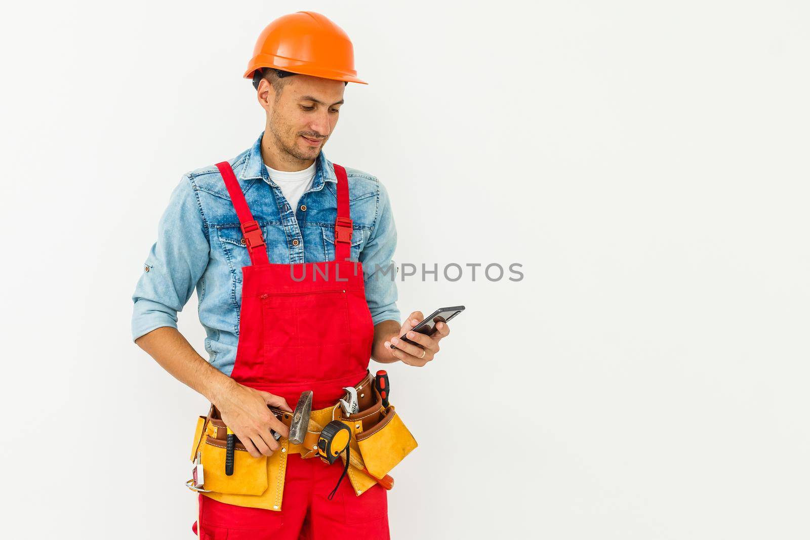 profession, construction and building - happy smiling male worker or builder in helmet and overall with smartphone over white background.