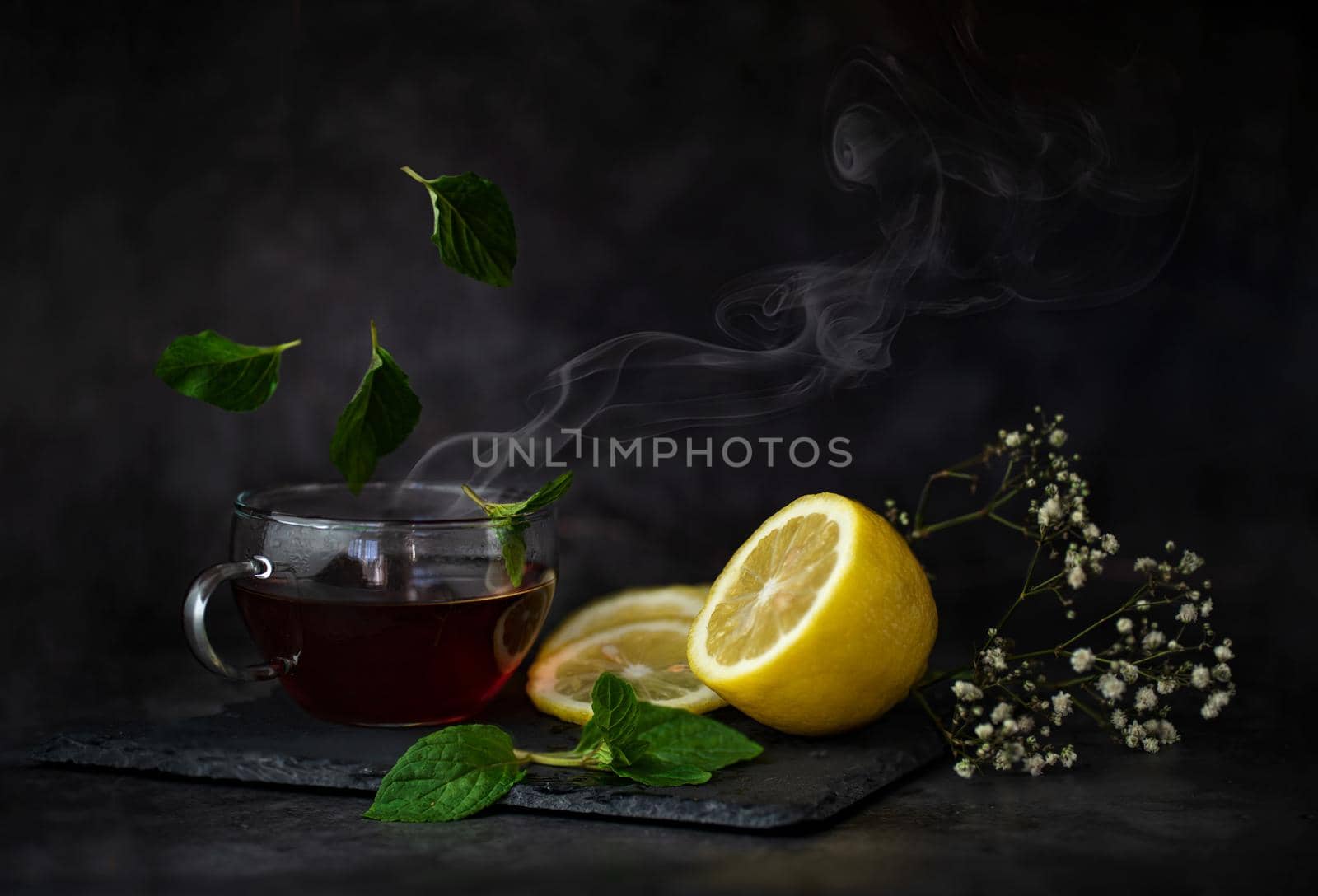 winter still life with a cut bright yellow lemon, a cup of fragrant tea and mint leaves floating around them on a table on a dark background High quality photo