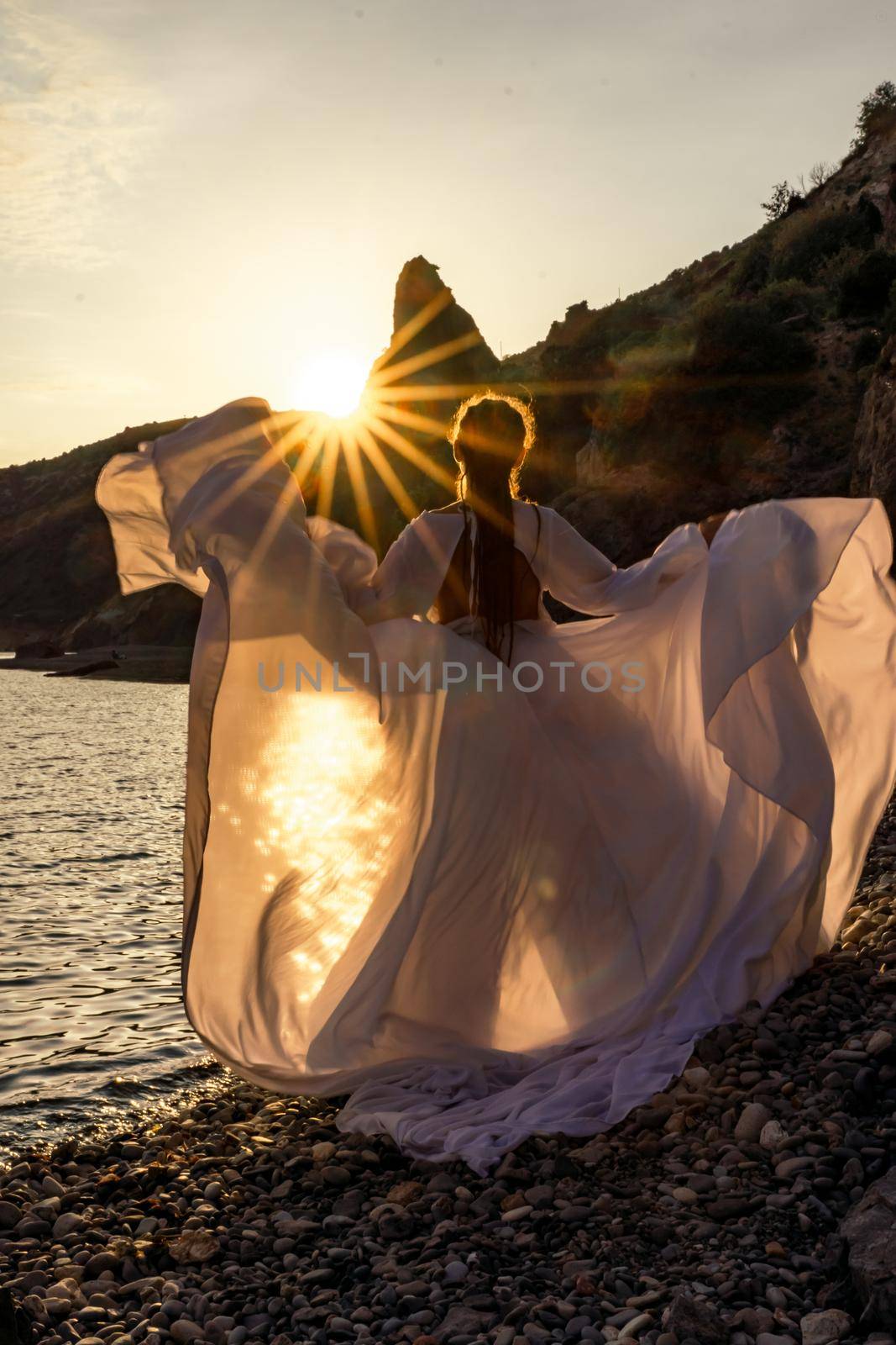 A mysterious female silhouette with long braids stands on the sea beach with mountain views, Sunset rays shine on a woman. Throws up a long white dress, a divine sunset