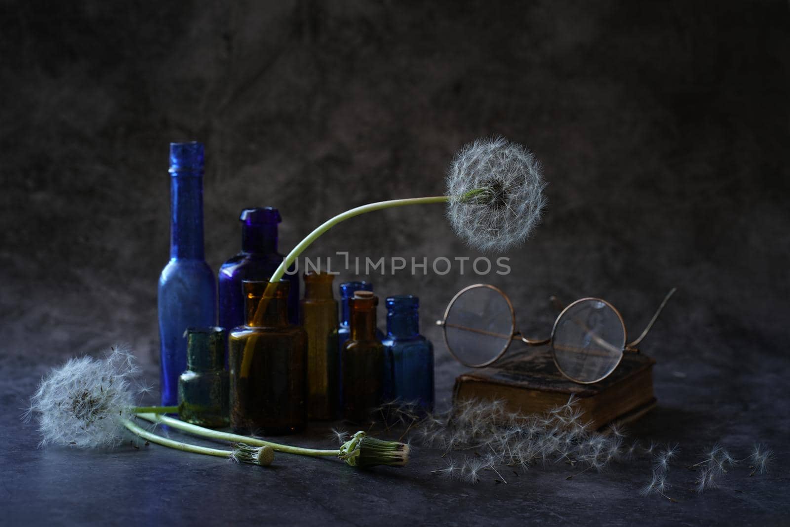 a still life consisting of dandelions with white fluffy heads multicolored small bottles and antique glasses of the late 19th and early 20th century on a table on a dark background. High quality photo