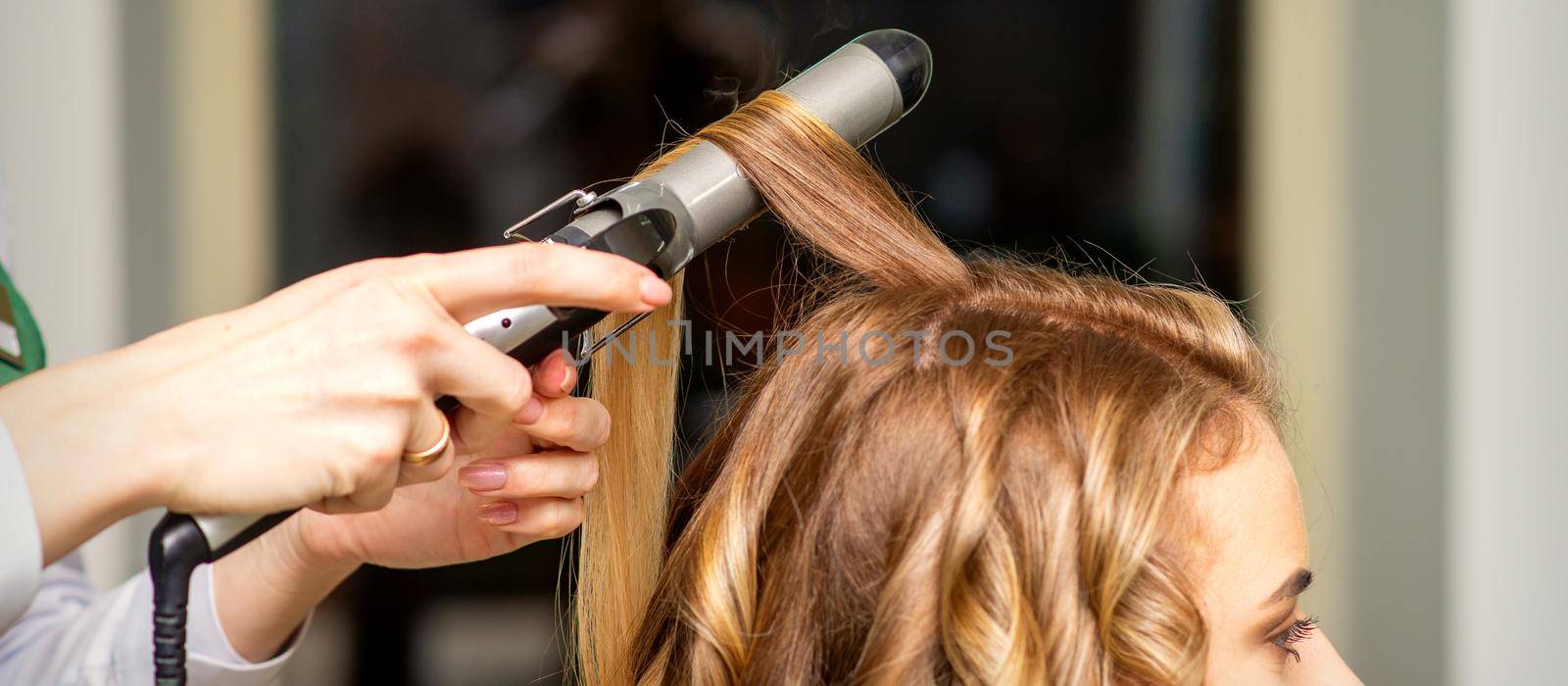 Hairdresser makes curls with a curling iron for the young woman with long brown hair in a beauty salon. by okskukuruza