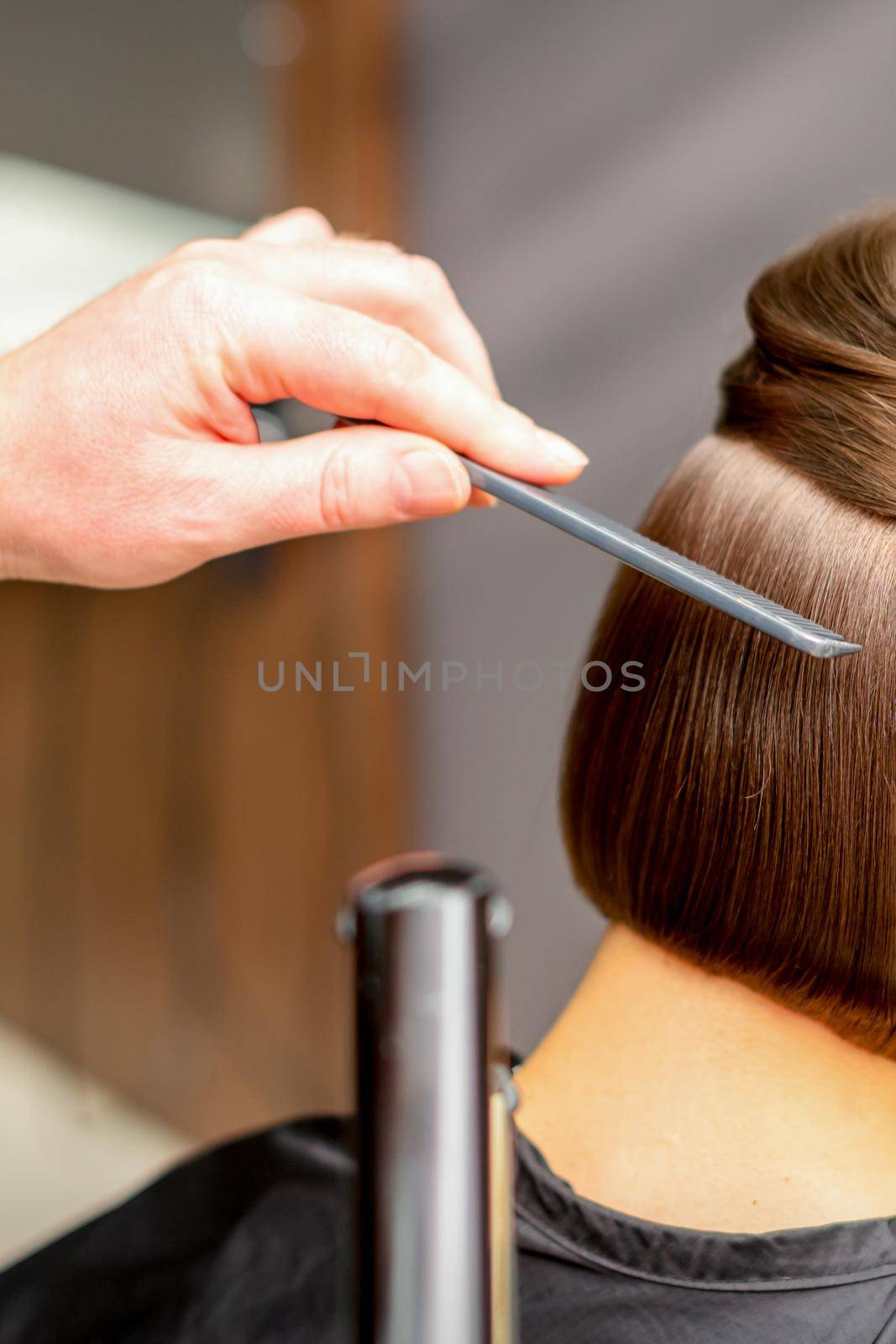 Hairstylist is straightening short hair of young brunette woman with a flat iron in a hairdresser salon, close up