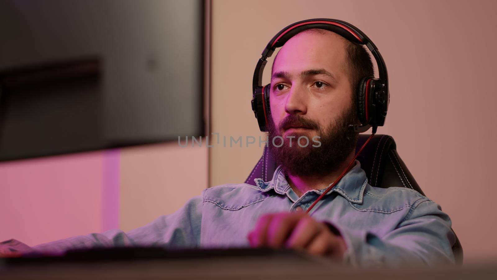 Handheld shot of man with gaming headset looking at computer screen and talking to team members while streaming gameplay. Gamer playing multiplayer online action game using professional pc setup.