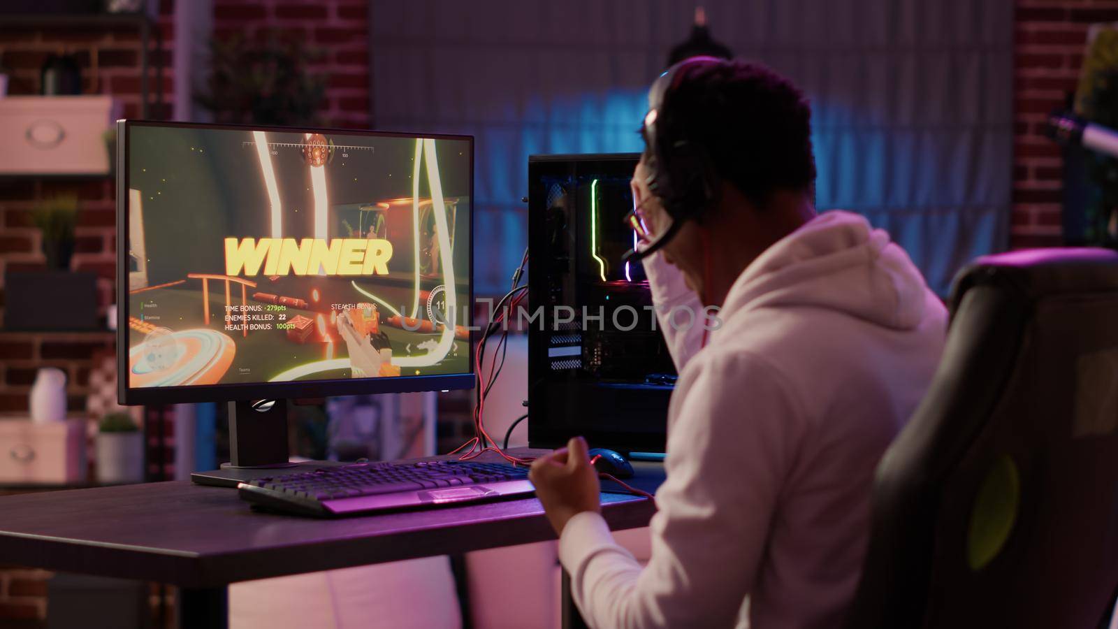 Over shoulder view of african american gamer celebrating after winning online competition on gaming pc. Man doing victory hand gesture happy after playing online multiplayer game first person shooter.