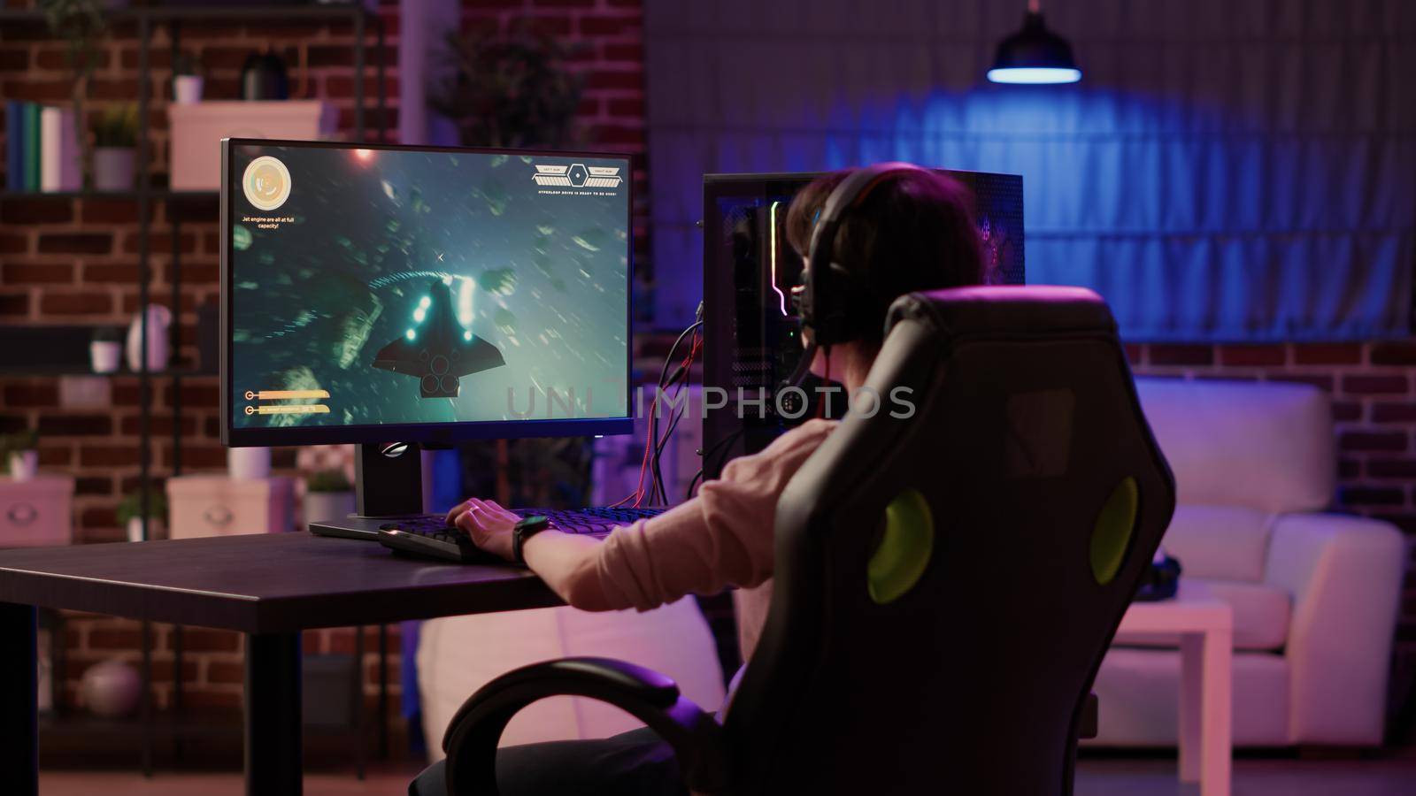 Over shoulder view of gamer girl relaxing and streaming on internet fast paced space shooter simulation gameplay while talking using headset. Woman playing rpg action game on professional pc setup.