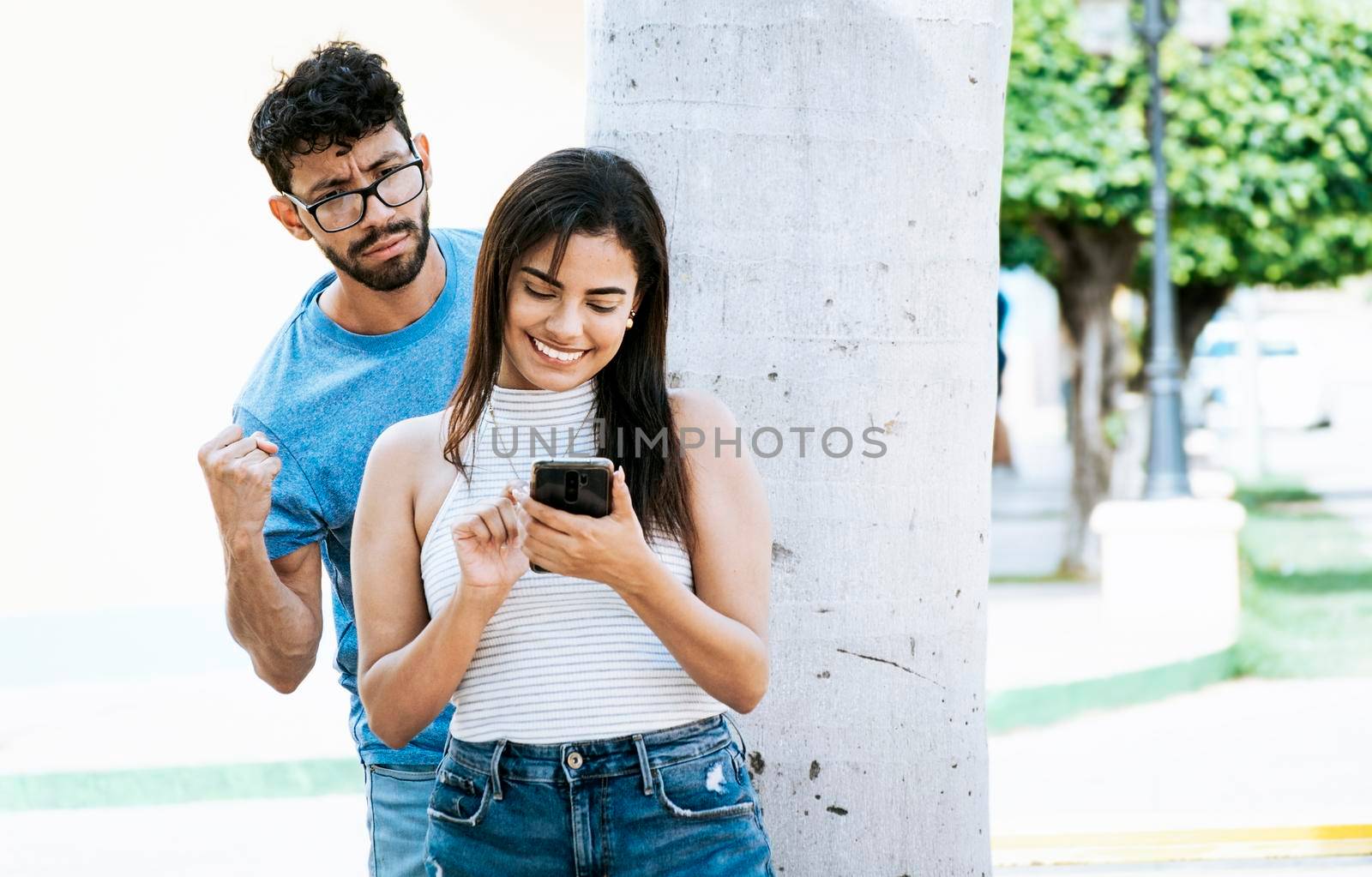 Jealous man spying on his girlfriend texting on the cell phone. Suspicious man spying on his girlfriend with his cell phone, Jealous boyfriend spying on his girlfriend's cell phone in the park by isaiphoto