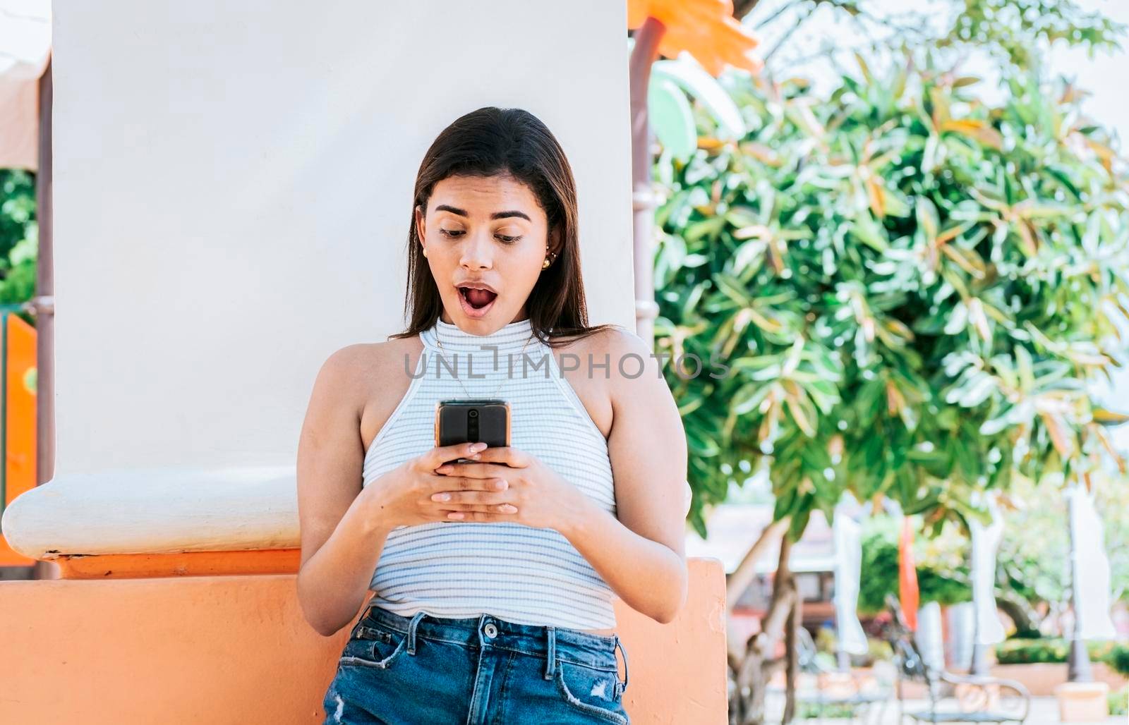 Surprised person looking at cell phone outdoor. Girl with cell phone with a surprised face leaning on a wall. Young woman with the cell phone with a gesture of surprise with open mouth outside by isaiphoto