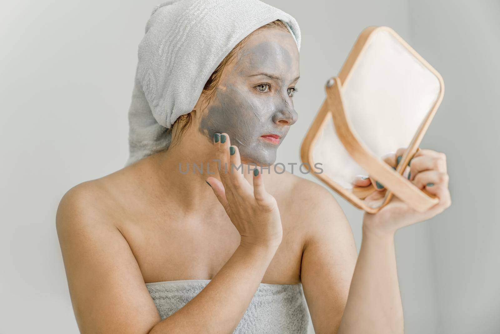 Young woman puts gray cosmetic clay mask on face while looking in mirror, her hair and body wrapped in towel, Side view. Close-up