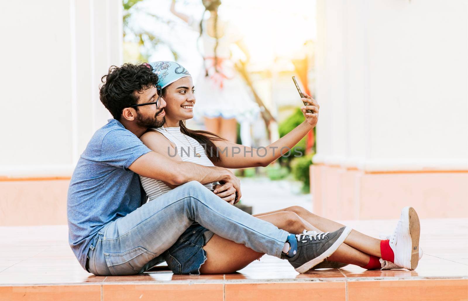 Young couple taking a selfie sitting on the floor. Young latin couple in love sitting on the floor taking a selfie outdoors. Concept of happy couple taking photos with the cell phone