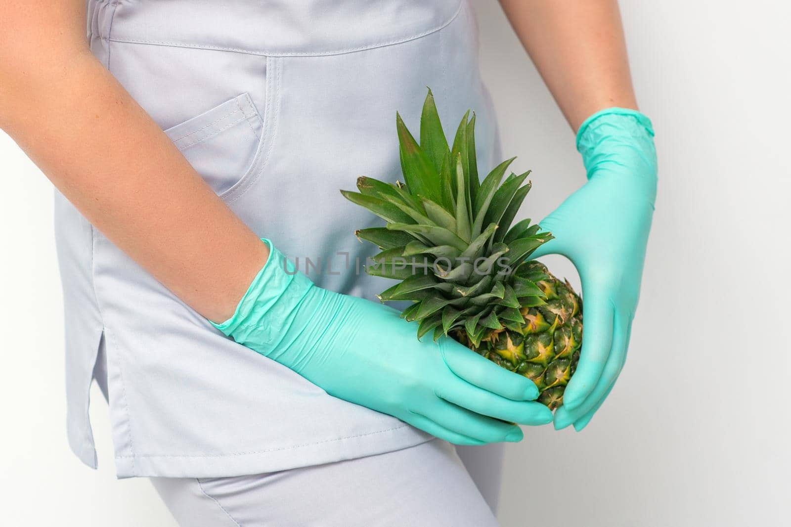 Young beautician wearing blue gloves in uniform with pineapple covers an intimate area on a white background, bikini zone depilation concept. by okskukuruza