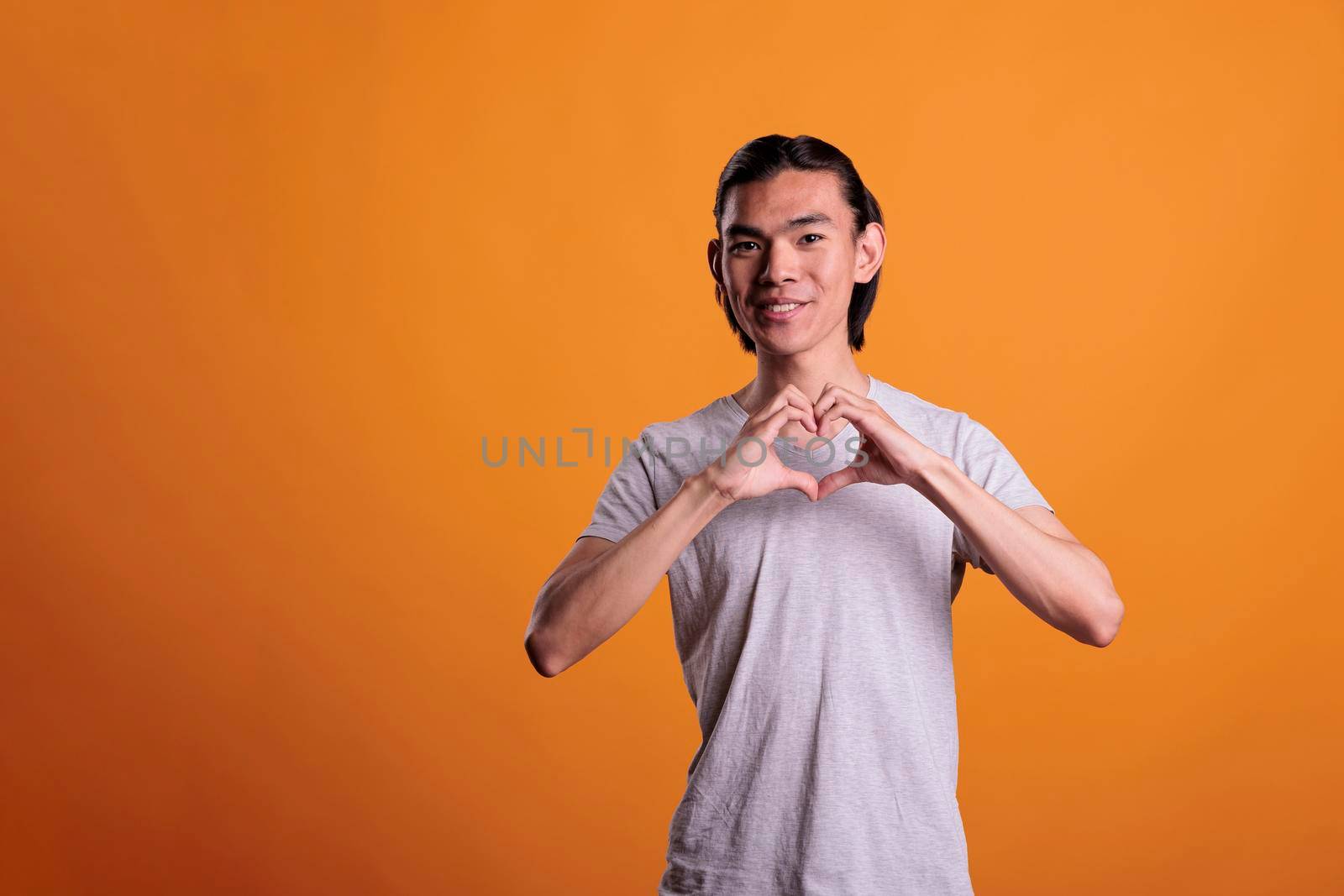 Young happy asian man showing heart shaped love gesture, romance symbol. Smiling cheerful teenager making appreciation sign, looking at camera with positive facial expression