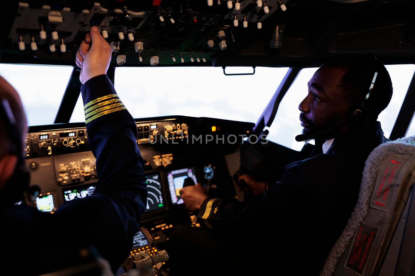 Multiethnic team of pilots using control panel in cockpit to fly airplane, pushing power switch and dashboard command buttons to takeoff. International airways service with aircrew in cabin.
