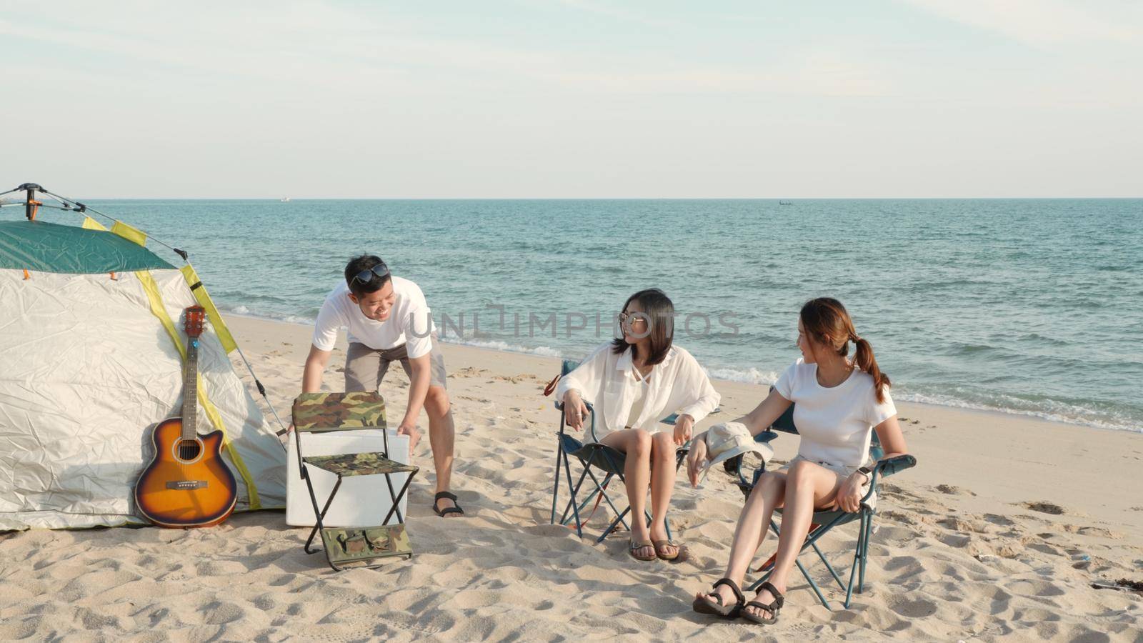 Happy friends with drinks in camp and laughing together in holidays on sand beach near camping tent vacation time at sunset by Sorapop