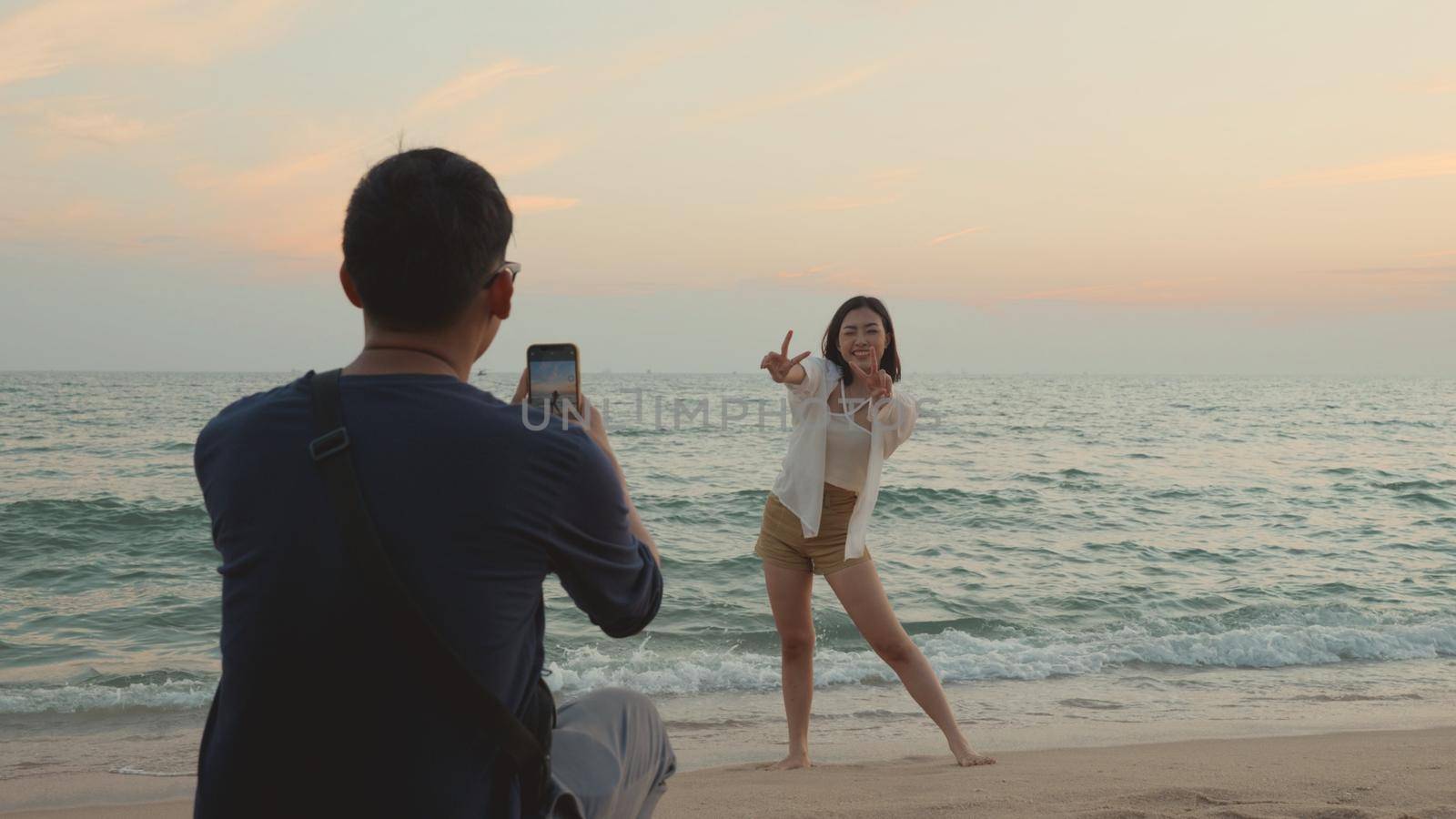 Happy man using smartphone camera making photo to woman at the beach and having fun together on beach by Sorapop