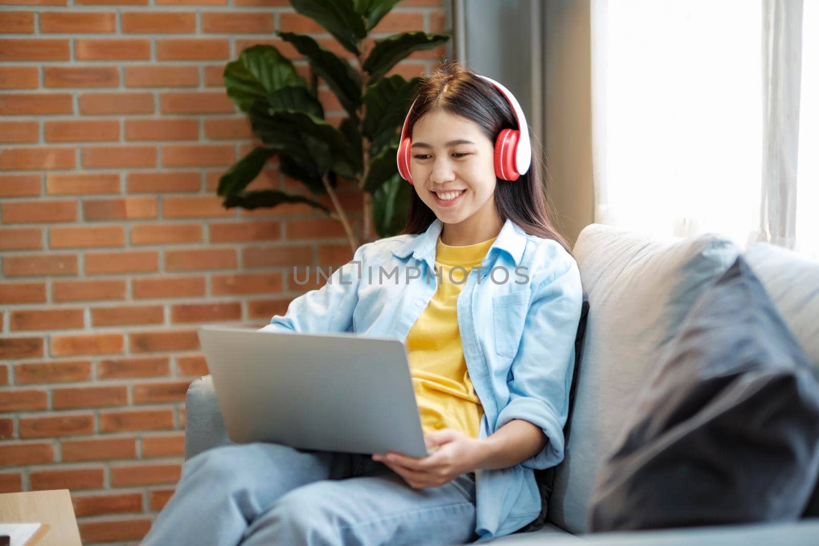 Young asian woman smiling listening to music using laptop while sitting on couch at home. by ijeab