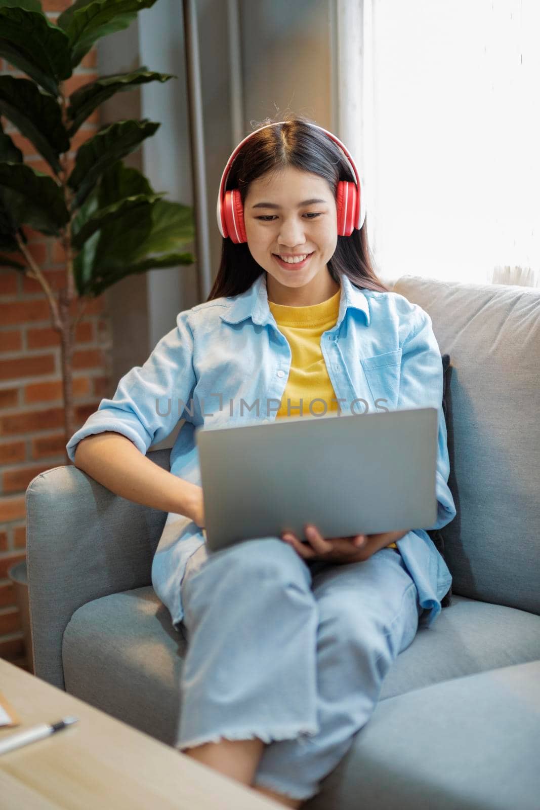Young asian woman studying, online learing, and listening to music with headset while sitting on couch at home. Listening to podcast, audio book, smiling, and, watching videos using laptop. 