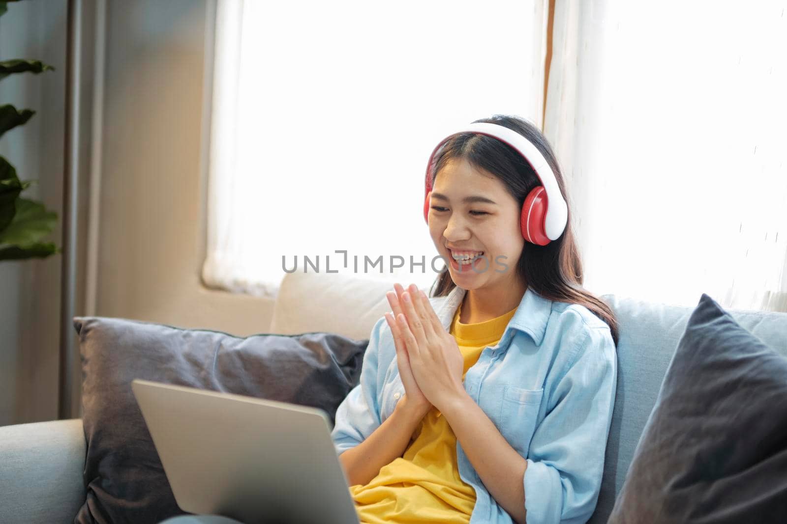 Happy young girl having video call on laptop with headset, studying online, communicating with friends, or working while sitting confortably on couch at home. Online learning and distant working concept.