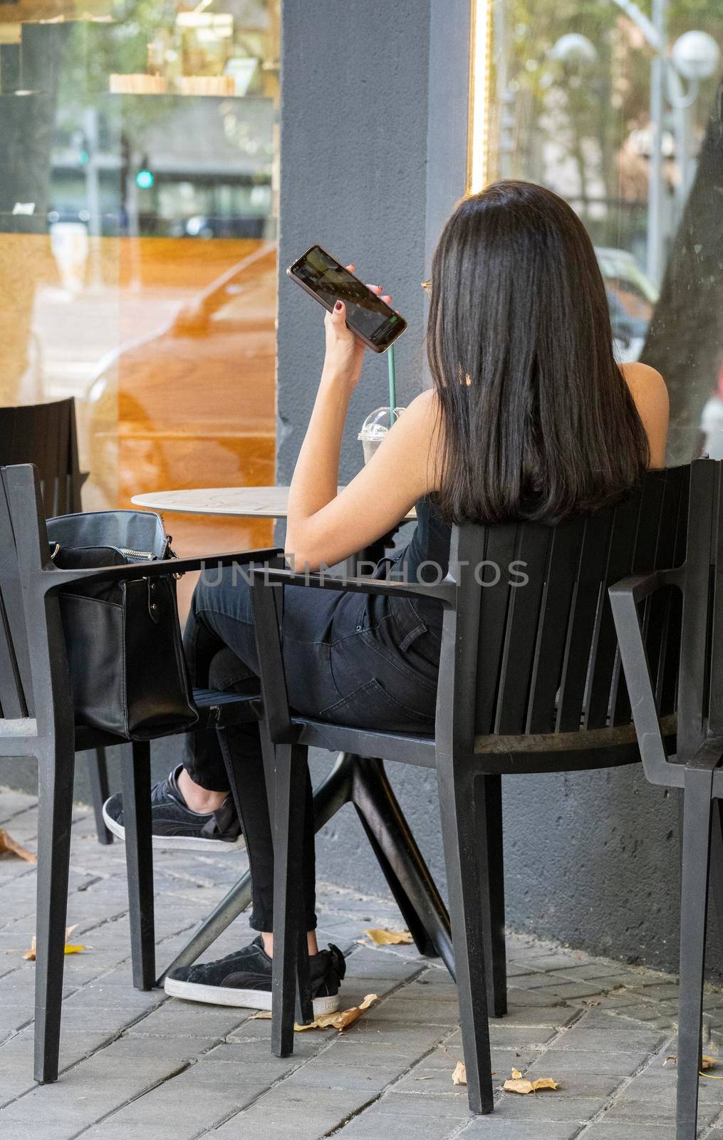 Young girl talking on phone from abroad with her family or friend, holding smartphone speaking, sitting at table outside a bar.