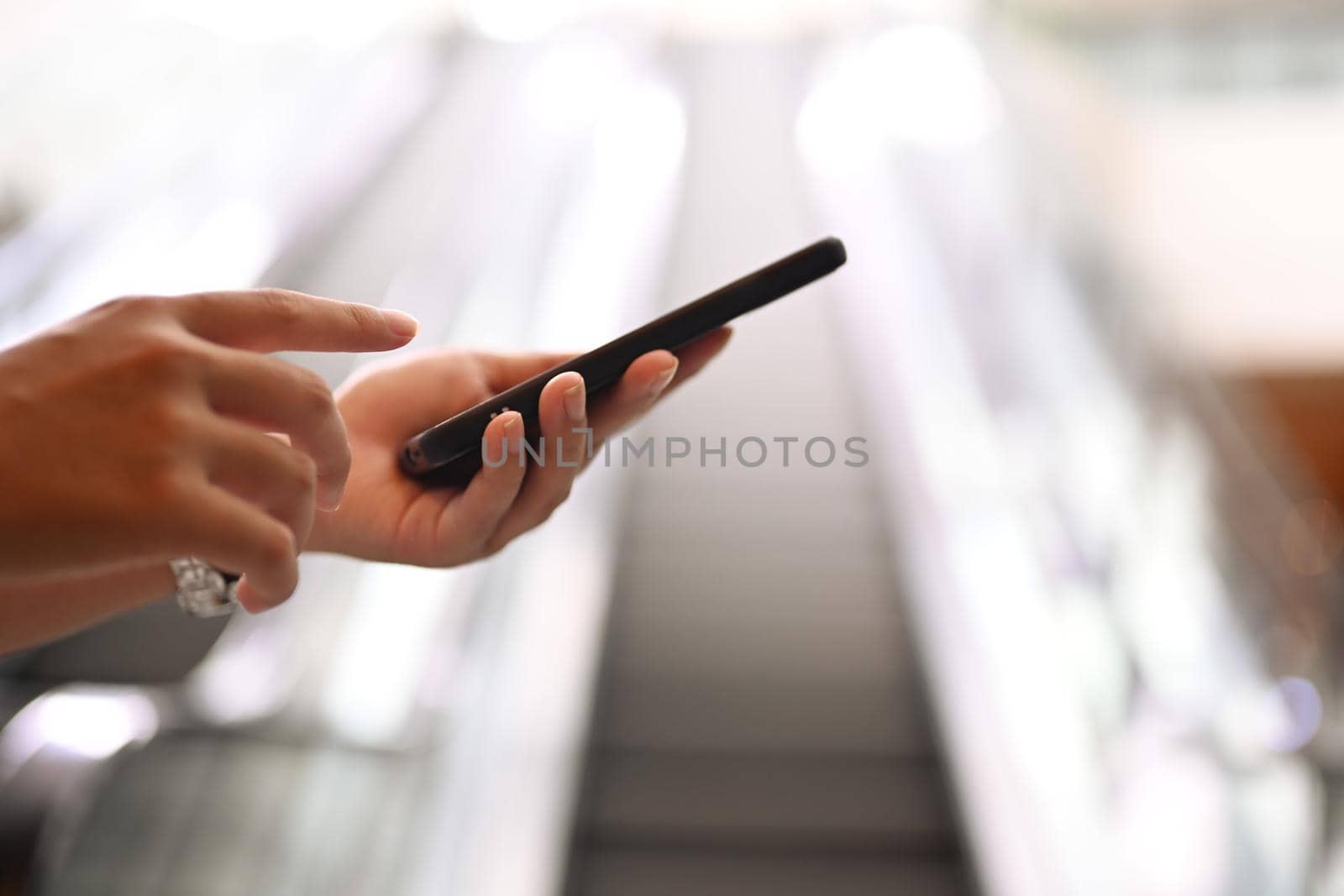 Woman hand typing on smartphone with blurred moving escalator and sunlight in background.