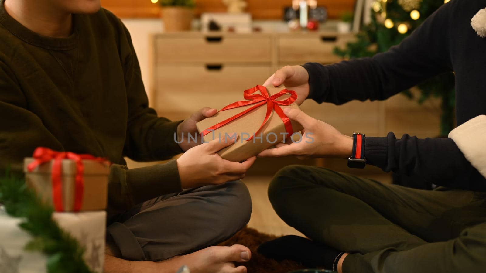 Image of friends exchanging presents while sitting near decorated Christmas tree in comfortable living room by prathanchorruangsak