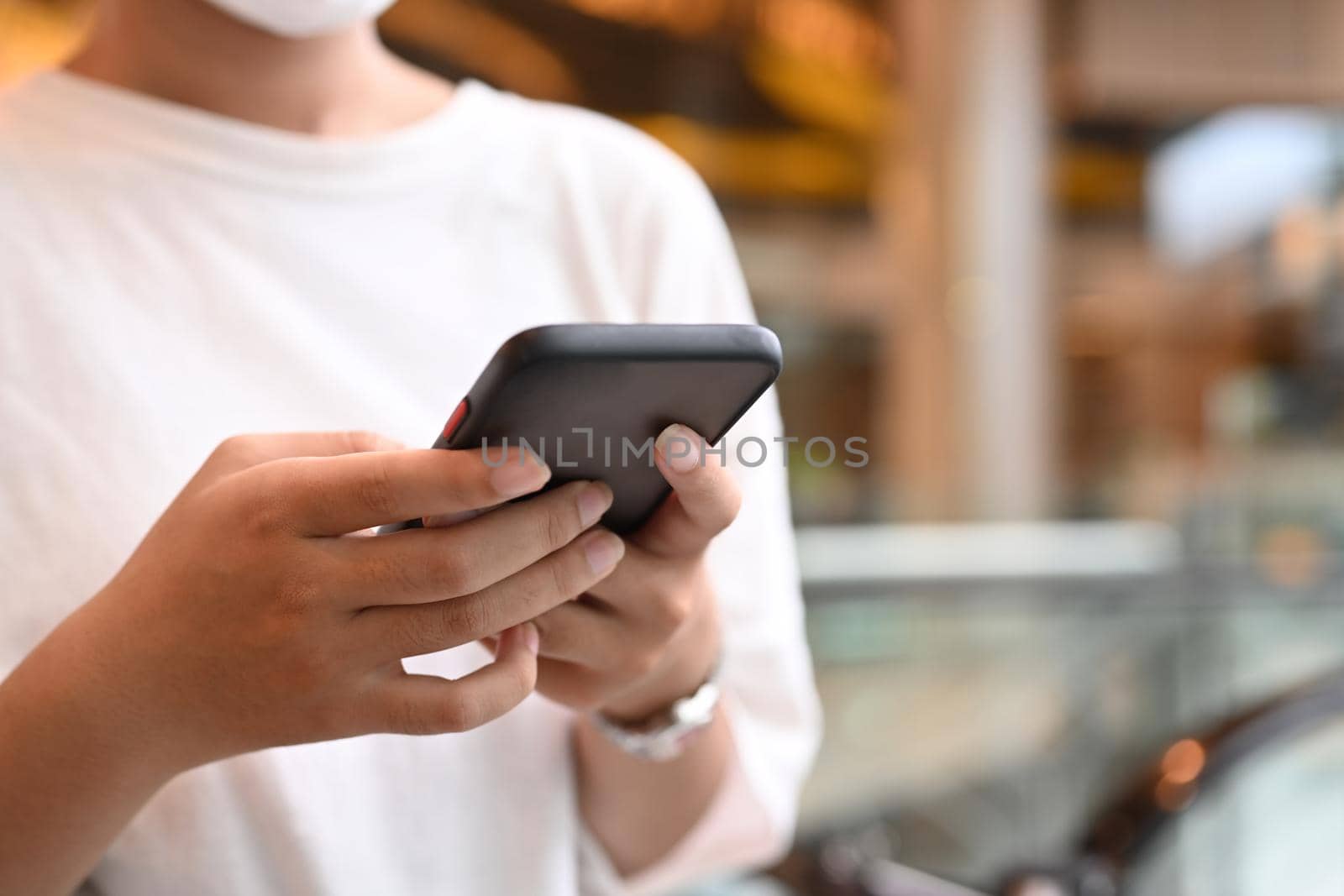 Woman hands typing message on smart phone, standing on blurred background of shopping mall and escalator.